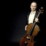 A photo of Gary Russell standing and holding a cello on a black backdrop.